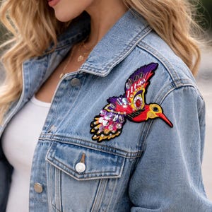 Puede incluir: Chaqueta vaquera con un parche de colibrí colorido. El parche presenta acentos rojos, morados, amarillos e iridiscentes. La chaqueta tiene un diseño clásico con cierres de botón y bolsillos.