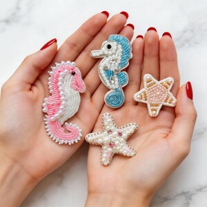 May include: A collection of beaded ocean-themed brooches, including two seahorses and two starfish. The seahorses are pink and blue, while the starfish are white and pink. Each brooch is intricately detailed with beads and pearls.