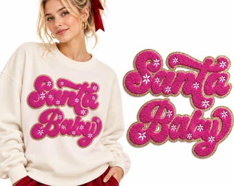 Puede incluir: Una sudadera de cuello redondo color crema con el texto Santa Baby en letras rosas texturizadas estilo felpa, con bordes de purpurina dorada y pequeños detalles de flores blancas. El diseño se muestra también por separado a la derecha.