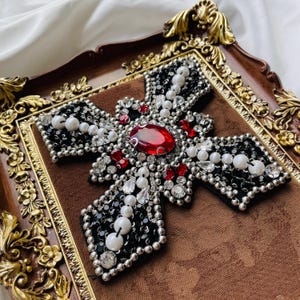 May include: A decorative cross with a large red gemstone at the center, surrounded by white pearls, red gems, and black beads. The cross is set on a brown fabric within a decorative, ornate frame with gold accents.