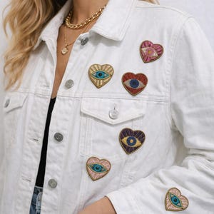 Puede incluir: Chaqueta vaquera blanca adornada con parches bordados en forma de corazón. Cada parche presenta un diseño de ojo con varias combinaciones de colores, incluyendo dorado, azul, rojo y rosa. La chaqueta tiene botones y bolsillos plateados.