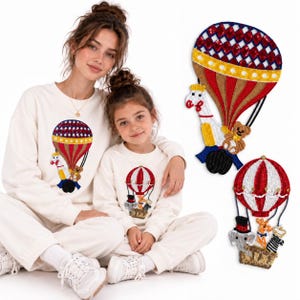 May include: Two people wearing matching cream-colored sweatshirts featuring a sequined hot air balloon graphic with a giraffe, elephant, and teddy bear. Two separate sequined hot air balloon patches are displayed to the right of the people.