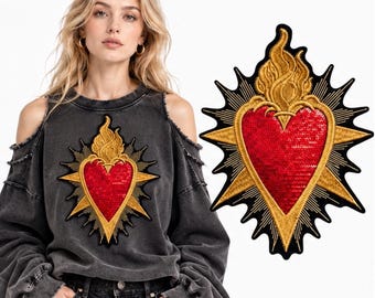 Puede incluir: Una sudadera gris carbón con hombros descubiertos que presenta un gran parche bordado de corazón sagrado. El parche muestra un corazón rojo de lentejuelas en el centro, rodeado de llamas doradas y rayos solares sobre un fondo negro.