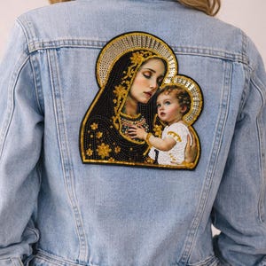 Puede incluir: Chaqueta vaquera azul claro con un parche de icono religioso en la espalda. El parche presenta una representación de la Virgen María y el niño Jesús, con adornos dorados y negros. La chaqueta tiene un diseño clásico.