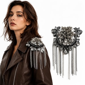 May include: A shoulder epaulet featuring a cluster of dark gray and clear faceted crystals arranged in a floral pattern, accented with a black fabric base and multiple dangling silver-toned metal chains that drape downward from the main piece.