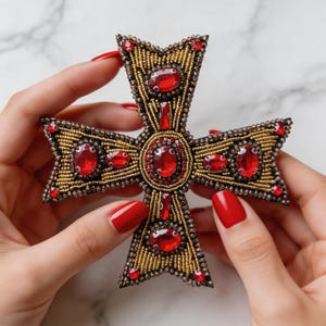 Puede incluir: Un broche detallado en forma de cruz. El broche presenta un diseño de cuentas doradas con grandes piedras preciosas rojas de forma ovalada y pequeños acentos rojos. La cruz tiene una apariencia texturizada y está sostenida por manos con esmalte de uñas rojo.
