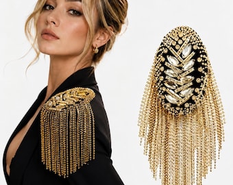 Puede incluir: Una charretera de hombro ovalada con una base de tela negra adornada con cristales transparentes de talla marquesa dispuestos en un patrón de hojas, rodeada de cuentas doradas y largos flecos de cadena de metal dorado en cascada.