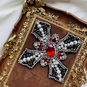 May include: A decorative cross with a large red gem at the center, surrounded by black and silver embellishments, white pearls, and red accents. The cross is set on a brown velvet background within a decorative, ornate wooden frame with gold accents.