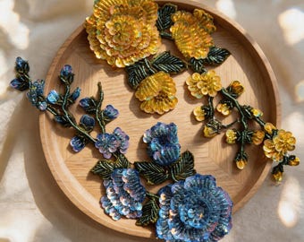 Puede incluir: Dos parches bordados florales con intrincados abalorios y lentejuelas. Un parche muestra flores amarillas y naranjas con hojas verde oscuro, mientras que el otro presenta flores azules y moradas con hojas verde oscuro, sobre una bandeja de madera.