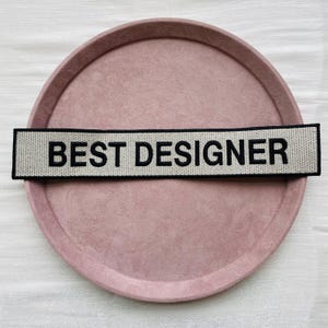 Puede incluir: Un parche rectangular con el texto "BEST DESIGNER" en letras negras sobre un fondo beige claro. El parche está colocado sobre una bandeja redonda de color rosa empolvado.