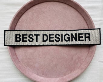 Puede incluir: Un parche rectangular con el texto "BEST DESIGNER" en letras negras sobre un fondo beige claro. El parche está colocado sobre una bandeja redonda de color rosa empolvado.