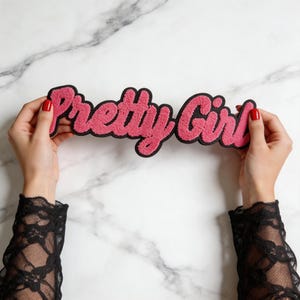 Puede incluir: Un parche de chenilla rosa con un borde negro que presenta el texto Pretty Girl en una fuente cursiva. El parche tiene una superficie texturizada y difusa, diseñado para aplicarse en artículos de tela como un adorno decorativo.