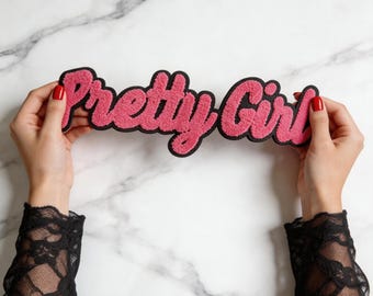 Puede incluir: Un parche de chenilla rosa con un borde negro que presenta el texto Pretty Girl en una fuente cursiva. El parche tiene una superficie texturizada y difusa, diseñado para aplicarse en artículos de tela como un adorno decorativo.