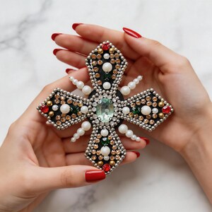 May include: A decorative cross-shaped brooch with a central green gemstone, surrounded by pearls, red, gold, and clear crystals. The brooch is held in a person's hands with red nail polish.
