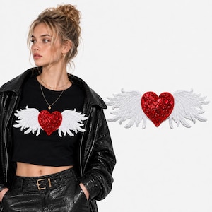 May include: A black crop top featuring a central red heart appliqué adorned with sparkling red sequins and white embroidered wings extending outward. The design is displayed on a black shirt and as a standalone patch on a white background.