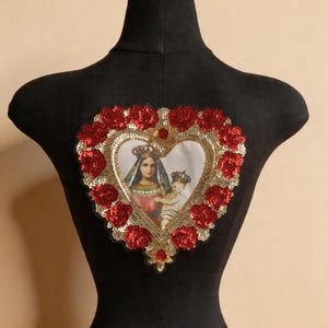 May include: A heart-shaped patch featuring an image of a woman and child, surrounded by red sequined roses and gold sequins. The patch is displayed on a black mannequin, showcasing its design and details.