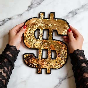 May include: A large dollar sign symbol patch covered in shimmering gold sequins. The symbol features a thick black fabric border outlining the shape, providing a distinct contrast against the metallic gold surface of the individual sequins.