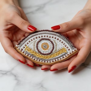 May include: A handcrafted, beaded eye-shaped brooch. The design features a white base with concentric circles in shades of gold, brown, and silver. The brooch is held by hands with red nail polish.