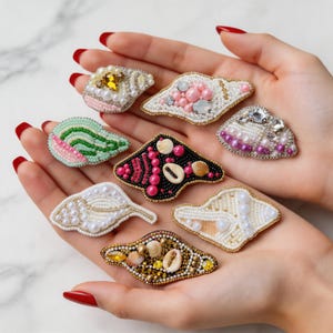 Op de afbeelding: Een verzameling handgemaakte, met kralen bezette schelpenbroches in verschillende kleuren en ontwerpen. Elke broche heeft ingewikkeld kralenwerk, parels en schelpen, met kleuren als roze, wit, goud en zwart. De broches worden op de handen van een persoon getoond.