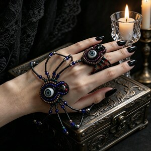 May include: A beaded hand ornament shaped like a spider, featuring two realistic glass eyes in shades of blue and grey. The spider body and legs are constructed from black, blue, and metallic beads, connecting a wrist bracelet to a finger ring.