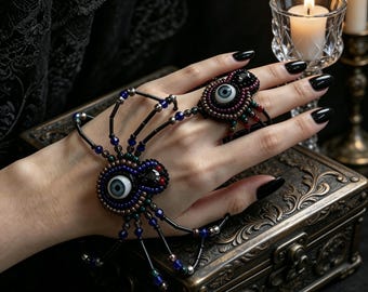 Könnte beinhalten: Ein perlenbesetzter Handschmuck in Spinnenform mit zwei realistischen Glasaugen in Blau- und Grautönen. Der Körper und die Beine der Spinne bestehen aus schwarzen, blauen und metallischen Perlen, die ein Armband mit einem Fingerring verbinden.