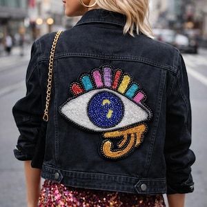 May include: Black denim jacket with a large, colorful beaded eye patch on the back. The eye design features a blue iris, white beaded sclera, and rainbow-colored lashes. A gold chain strap hangs from the shoulder.