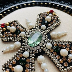 May include: Close-up of a decorative cross with a black velvet base. The cross is embellished with a large, light green gemstone at the center, surrounded by smaller clear and amber-colored crystals, pearls, and silver beads. Red and green accents are also present.