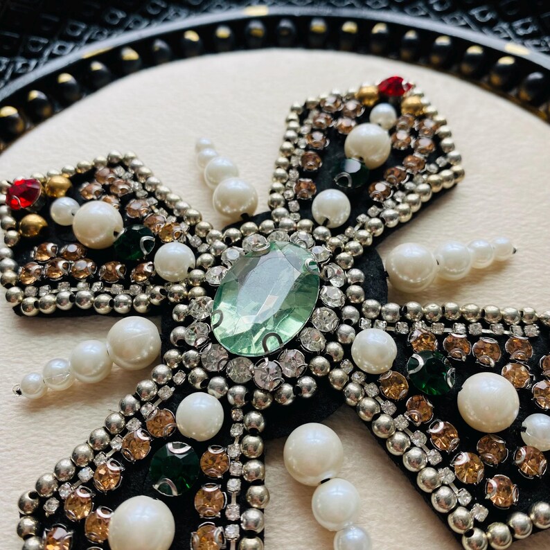 May include: Close-up of a decorative cross with a black velvet base. The cross is embellished with a large, light green gemstone at the center, surrounded by smaller clear and amber-colored crystals, pearls, and silver beads. Red and green accents are also present.