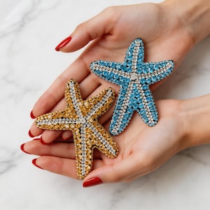 May include: Two bejeweled starfish, one gold and one blue, are held in open hands. The starfish are covered in sparkling crystals, with a silver border. The background is a light, neutral color.