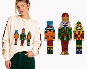 Puede incluir: Una sudadera de manga larga color crema con tres diseños de cascanueces de lentejuelas en la parte delantera y motivos adicionales de cascanueces y copos de nieve de lentejuelas en las mangas, en tonos rojo, verde, azul y dorado.