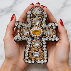 May include: A black cross-shaped brooch, heavily embellished with clear and amber-colored crystals and gold-colored beads. The brooch has a central oval amber stone and is held in a person's hands.