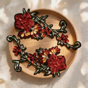 Könnte beinhalten: Zwei florale Stickaufnäher mit dunkelroten Stoffblumen, grünen Ranken und glitzernden goldenen sowie roten Pailletten. Die Aufnäher haben einen schwarzen Rand und sind auf einem runden Holztablett angeordnet.