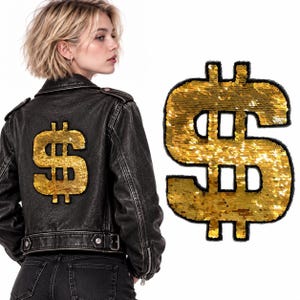 May include: A black leather jacket featuring a large, gold sequined dollar sign patch on the back. A separate, matching gold sequined dollar sign patch is displayed next to the jacket, showcasing the reflective, metallic texture of the sequins.