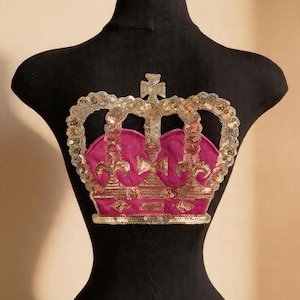 Pode incluir: Aplique decorativo de coroa com centro rosa e acabamento em lantejoulas douradas, exibido em um manequim preto. A coroa apresenta uma cruz no topo e é adornada com lantejoulas. O manequim tem um pescoço dourado.