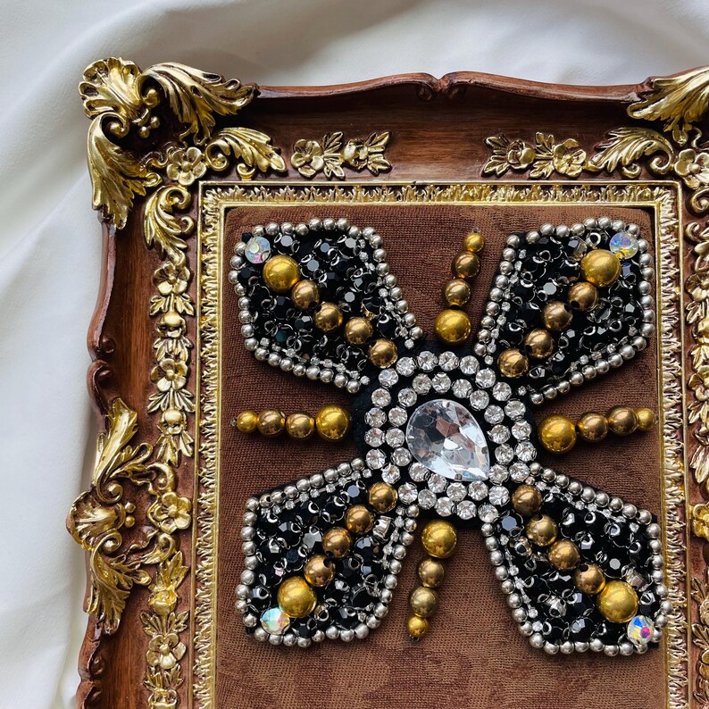May include: A decorative butterfly design, featuring black beads, gold accents, and a large clear gem, set within a brown velvet background. The butterfly is framed by a gold-colored border and a dark brown, ornate wooden frame.