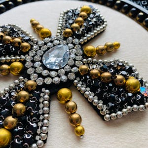 May include: A decorative brooch in the shape of a bee, featuring a large clear crystal center stone, surrounded by small clear crystals. The wings are black with silver and black beads, and gold-colored spherical beads are used throughout the design.