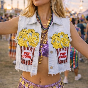 May include: A light-wash denim vest with two popcorn-themed patches. The patches feature yellow sequined popcorn in a red and white popcorn container. The outfit includes a purple sequined top and shorts, and a gold coin belt.