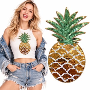 May include: A white sleeveless crop top featuring a large pineapple applique made of shiny gold and green sequins. The pineapple design has a textured, scale-like pattern in gold for the fruit body and vibrant green for the leafy top section.