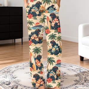May include: Cream-colored pajama pants featuring a repeating pattern of dark blue monsters wearing floral shirts and sunglasses. The print includes tropical elements like palm trees, red hibiscus flowers, surfboards, coconuts with straws, and a sunset over the ocean.