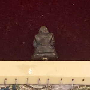 May include: A small, dark-colored figurine of a seated figure, possibly a Buddha or similar religious icon. The figure is seen from the back, with a dark maroon background. The figurine is on a light-colored surface with small holes.