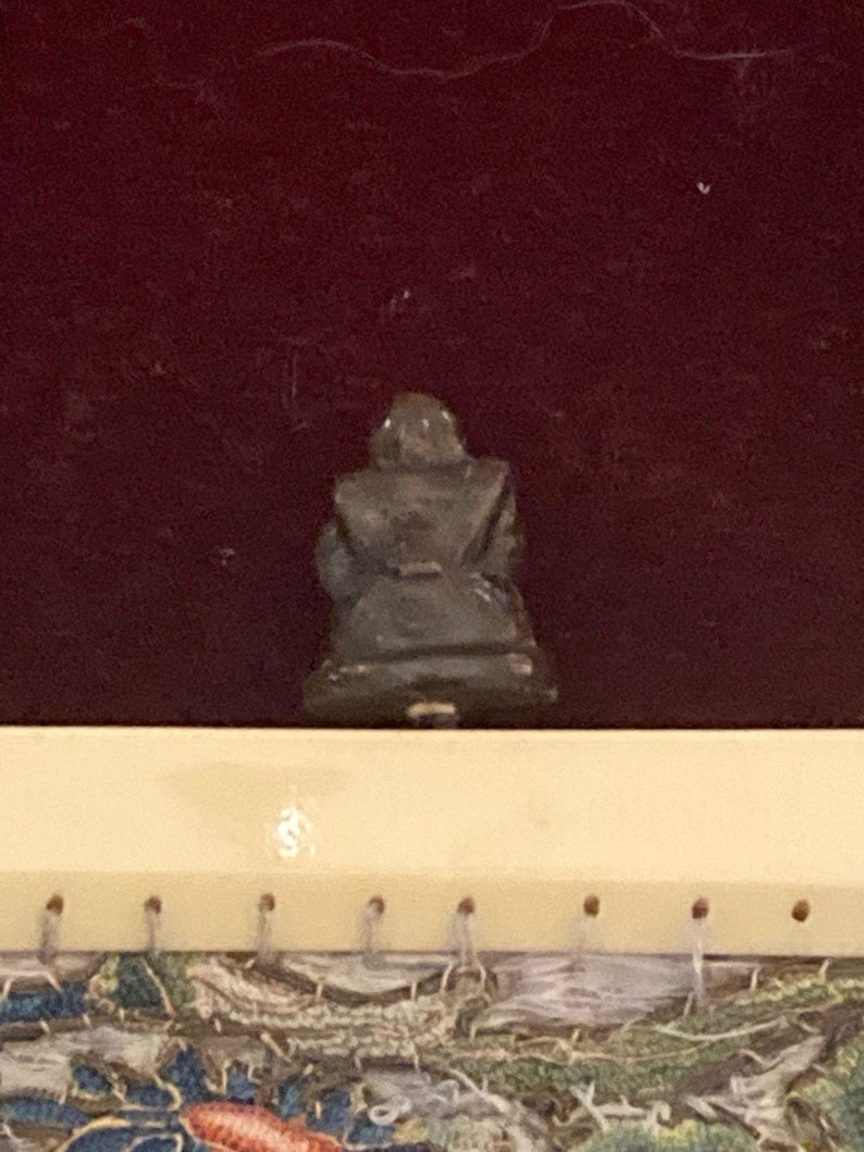 May include: A small, dark-colored figurine of a seated figure, possibly a Buddha or similar religious icon. The figure is seen from the back, with a dark maroon background. The figurine is on a light-colored surface with small holes.