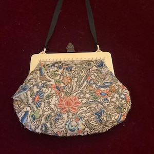 May include: An antique beaded handbag with a cream-colored frame and a black handle. The bag features a floral design with pink, blue, and green beads on a white background. The bag is likely from the early 20th century.