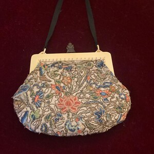 May include: An antique beaded handbag with a cream-colored frame and a black handle. The bag features a floral design with pink, blue, and green beads on a white background. The bag is likely from the early 20th century.