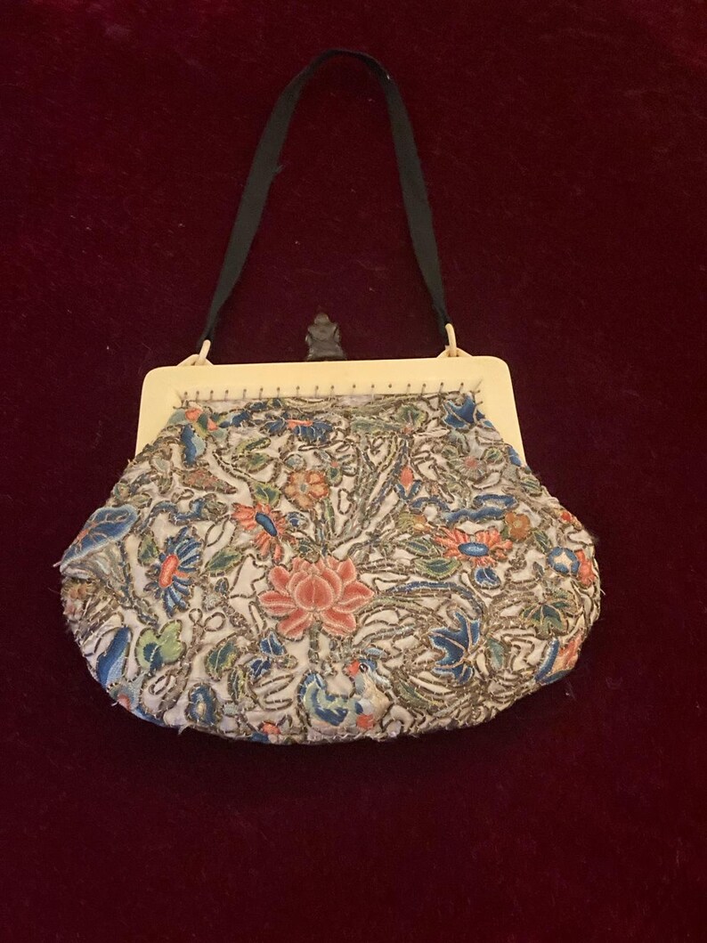 May include: An antique beaded handbag with a cream-colored frame and a black handle. The bag features a floral design with pink, blue, and green beads on a white background. The bag is likely from the early 20th century.