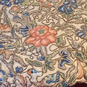 May include: Close-up of an embroidered textile with a floral design. The embroidery features a variety of colors, including pink, blue, green, and orange, on a cream-colored background. The top edge is adorned with turquoise and pink beads.
