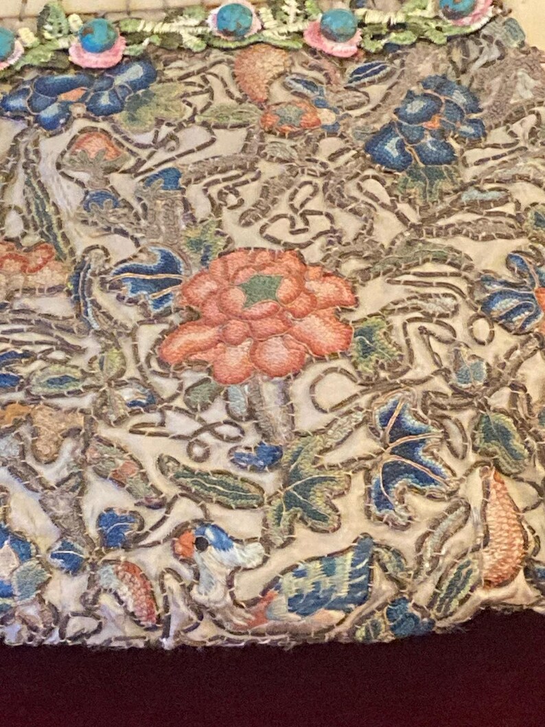 May include: Close-up of an embroidered textile with a floral design. The embroidery features a variety of colors, including pink, blue, green, and orange, on a cream-colored background. The top edge is adorned with turquoise and pink beads.