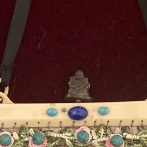May include: A small, dark figurine of a seated Buddha is centered above a decorative purse. The purse features turquoise and blue gemstones, and embroidered floral patterns in pink, blue, and green.