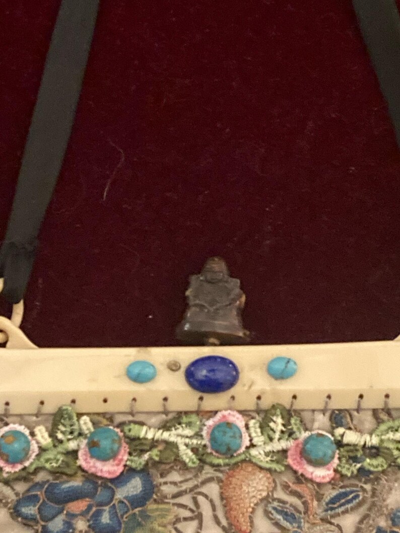 May include: A small, dark figurine of a seated Buddha is centered above a decorative purse. The purse features turquoise and blue gemstones, and embroidered floral patterns in pink, blue, and green.
