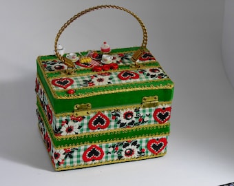 Puede incluir: Una caja decorativa con asa y detalles dorados. La caja es verde con un estampado de cuadros y diseños de corazones y flores rojos. Un juego de té en miniatura y pasteles están en la parte superior. La caja tiene un cierre y ribete dorados.