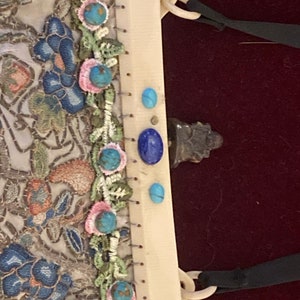 May include: Ivory-colored vintage handbag with intricate floral embroidery in blue, peach, and green. Embellished with turquoise and lapis lazuli stones. Features a cream-colored handle and black ribbon detail.